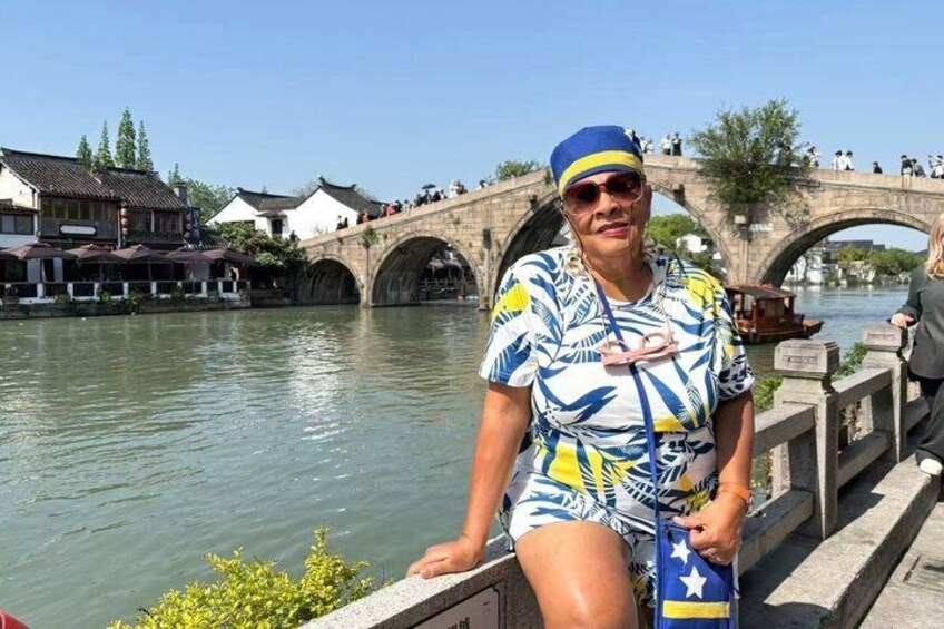 Une personne vêtue d’une tenue bleue et blanche se tient au bord d’une rivière, avec un pont et des bâtiments traditionnels en arrière-plan. 5