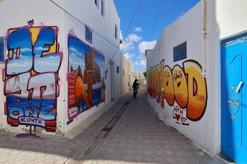A narrow street with colorful graffiti on the walls, a blue door, and a clear sky. 3