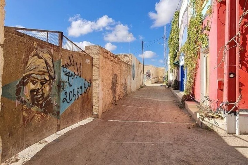 A narrow alley with a mural on the left wall, a red building on the right, and a clear blue sky above. 2