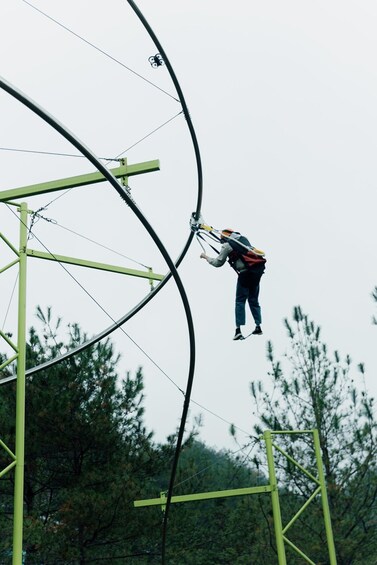 A person suspended on a cable, wearing a harness and helmet, against a backdrop of trees and utility poles. 3