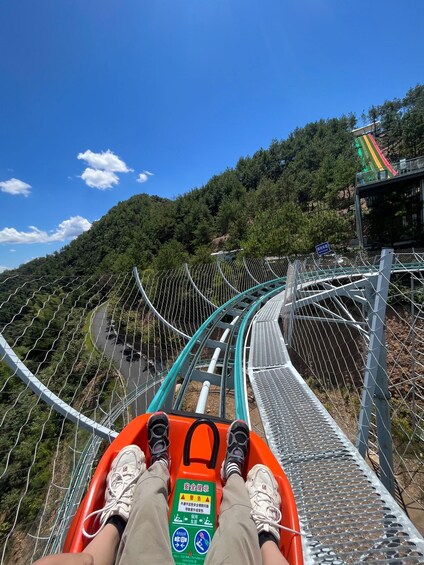 A person seated in a safety-deployed ride vehicle on a metal track, surrounded by a forested hillside and a colorful building structure. 2