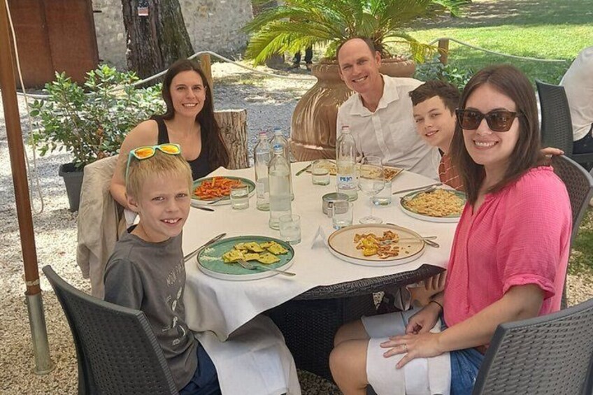 En familie spiser udendørs ved et rundt bord med tallerkener mad, omgivet af stole og en have. 4