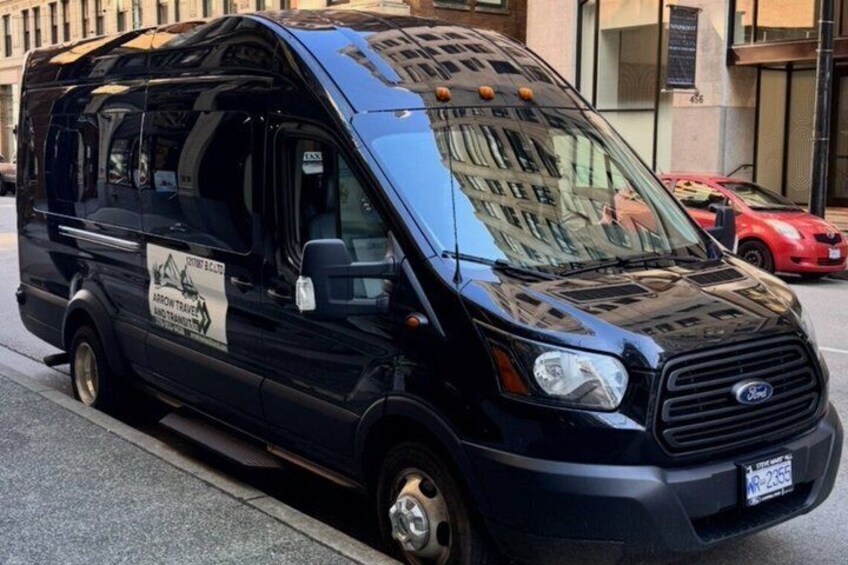 A black Ford Transit van parked on a city street with a 'Taxi' sign on the windshield and a 'Taxi' sign on the side. 2