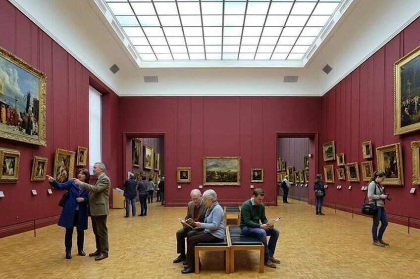 Une galerie d’art aux murs rouges et au plancher en bois, où l’on peut voir plusieurs personnes contempler des tableaux. 2