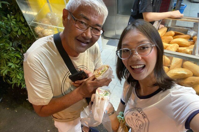Two people holding food items in a food stall area with bread displayed in the background. 5
