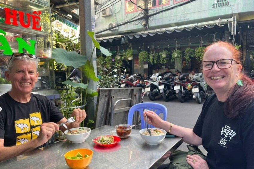 Dos personas cenando en una mesa al aire libre con tazones de comida, una silla azul y una calle con motocicletas estacionadas al fondo. 4