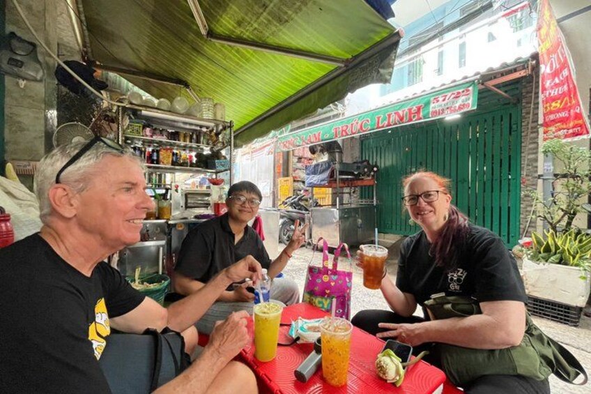 Tres personas sentadas en una mesa al aire libre con bebidas y aperitivos, un toldo verde y una tienda en el fondo. 5