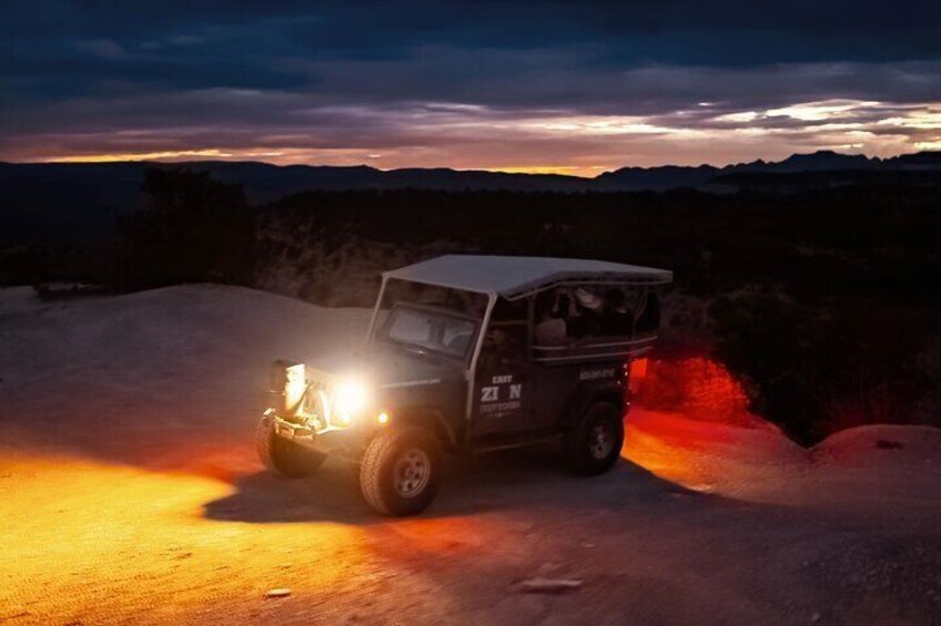A vehicle with headlights on, parked on a dirt road with a sunset in the background. 3