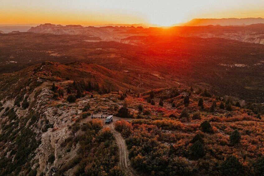 A mountainous landscape at sunset with a vehicle and a small building on a dirt path. 1