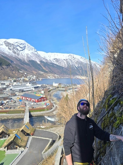 A person standing on a cliff overlooking a valley with a river, buildings, and snow-capped mountains. 1