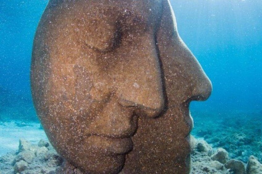A large, weathered head sculpture partially submerged in clear blue water. 4