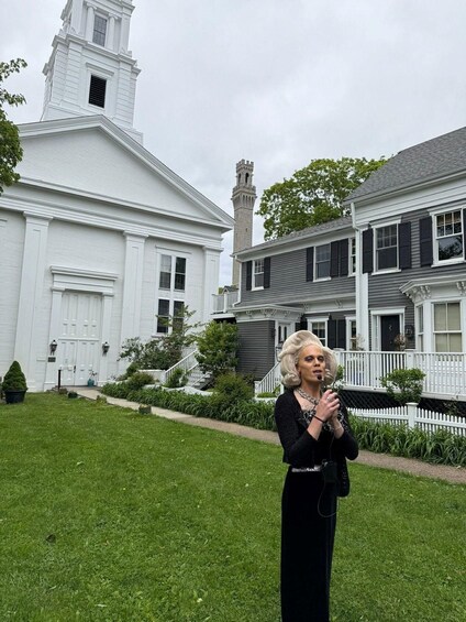 A person standing in front of a white church with a steeple and a grey house with black shutters. 2