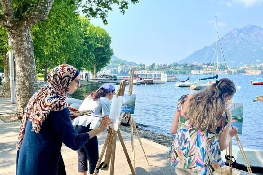 Three people painting by a lakeside with boats and mountains in the background. 1