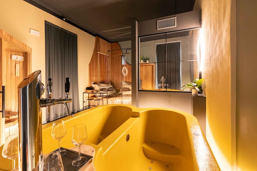 A yellow bathtub with two empty wine glasses, a wooden sauna door, and a mirror reflecting a plant. 4