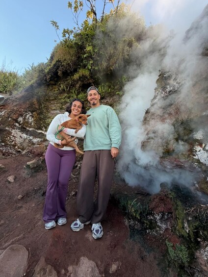 Two people standing on a rocky terrain with steam rising from the ground, surrounded by greenery and a clear sky. 2