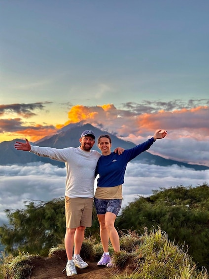 Two people standing with arms outstretched on a mountain trail with a sunset and clouds in the background. 1