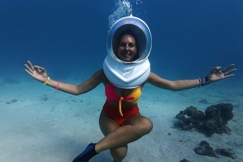 Eine Person trägt Unterwasserhelm, umgeben von klarem, blauem Wasser und sandigem Meeresboden. 1