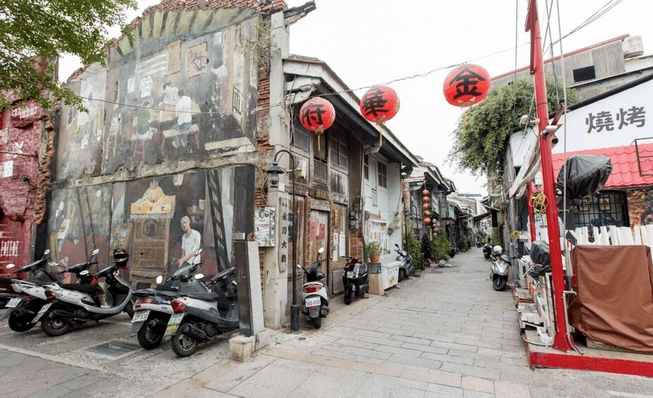 A narrow street lined with traditional buildings, some with visible signs and decorations, and parked scooters. 5