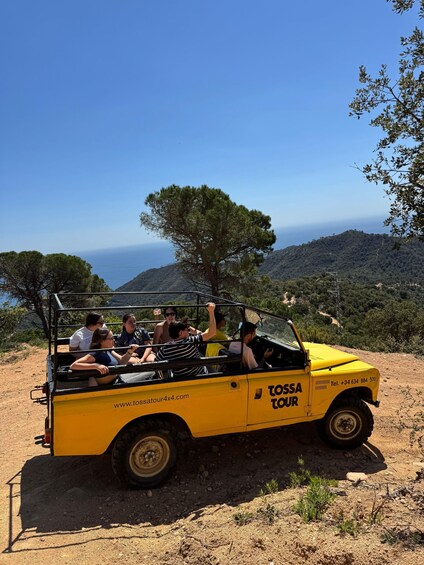 A yellow off-road vehicle with passengers, parked on a dirt road with a scenic view of hills and a clear sky. 1