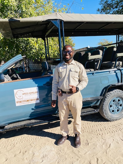 A man standing next to a safari vehicle with a canopy, in a sandy area with trees in the background. 1