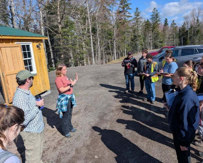A group of people standing outdoors near a wooden structure and a parked vehicle. 3
