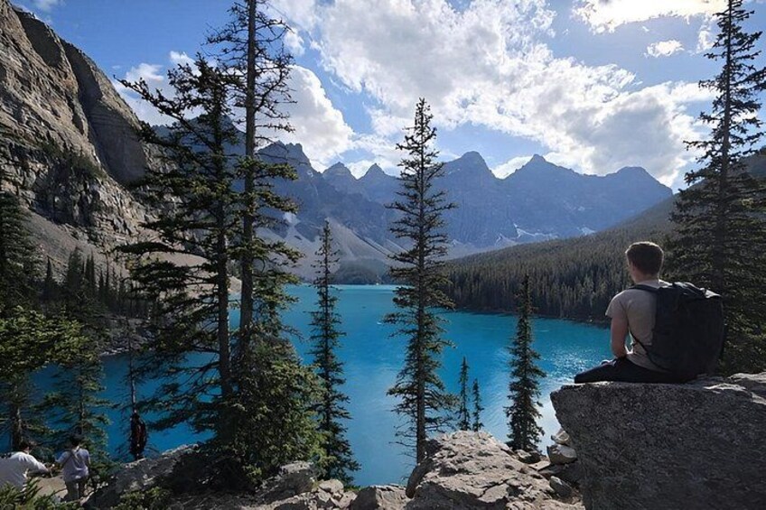A person sitting on a rock by a turquoise lake surrounded by mountains and pine trees. 5