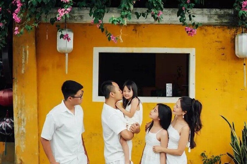 A family of five in front of a yellow building with a window and hanging flower pots. 2