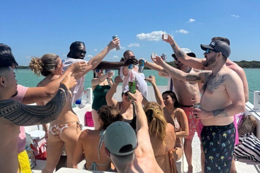 Un grupo de personas en un bote, algunas sosteniendo bebidas, con un cuerpo de agua y un cielo despejado al fondo. 2