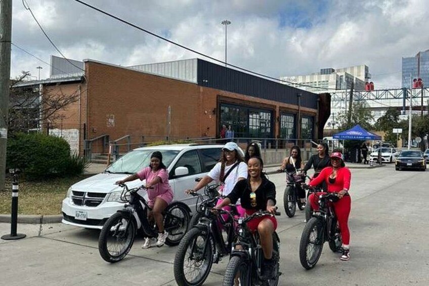 Un gruppo di persone in bicicletta in un contesto urbano, con un edificio e auto parcheggiate sullo sfondo. 2