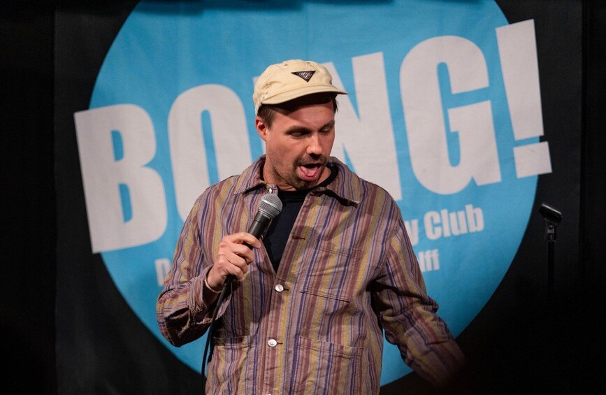 A person holding a microphone in front of a sign with 'BOING!' and 'Comedy Club' text. 5