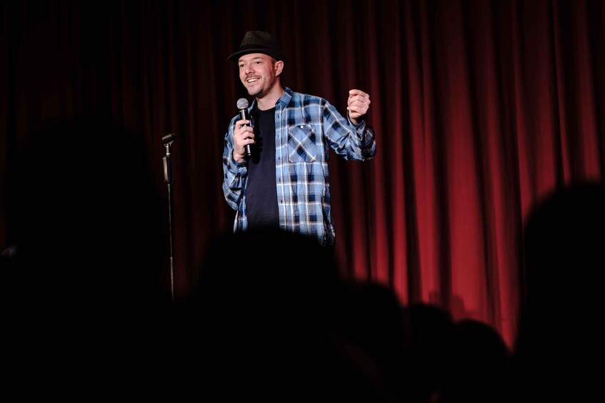 A person on stage with a microphone, wearing a plaid shirt and a hat, in front of a red curtain backdrop. 2