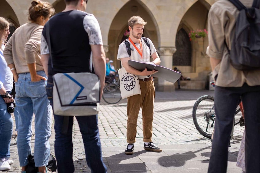 A person holding a map and a bag with a logo, standing in a courtyard with arches and a bicycle. 3