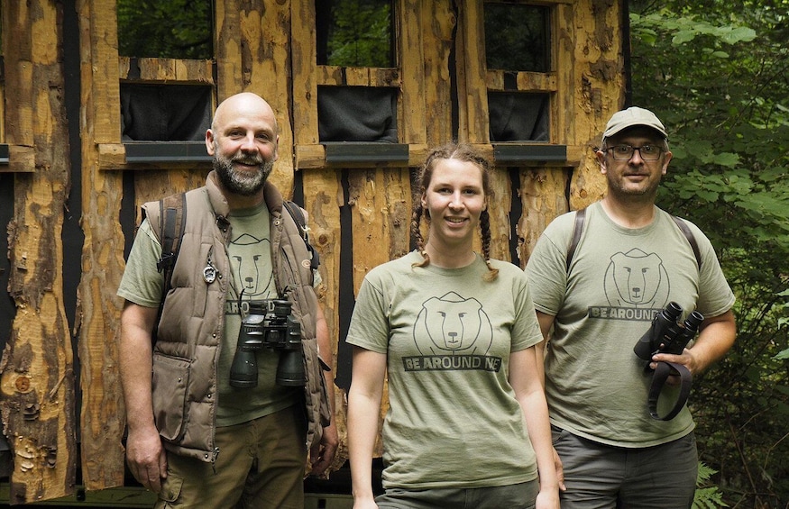 Three people standing in front of a wooden structure with multiple windows. 2
