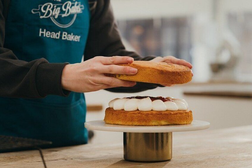 A person wearing a blue apron labeled 'Head Baker' holding a piece of bread above a cake on a white plate. 4