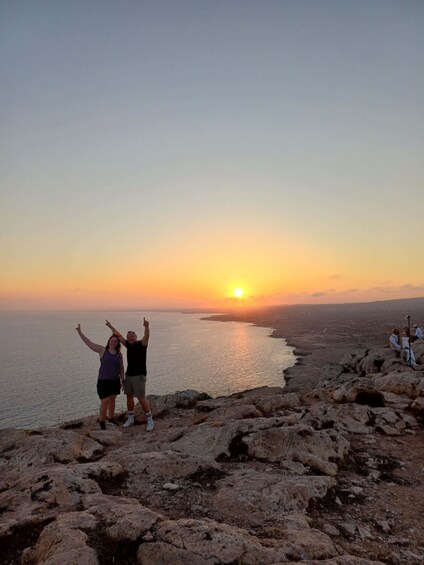 A sunset over a rocky coastline with two people posing with their arms raised. 1