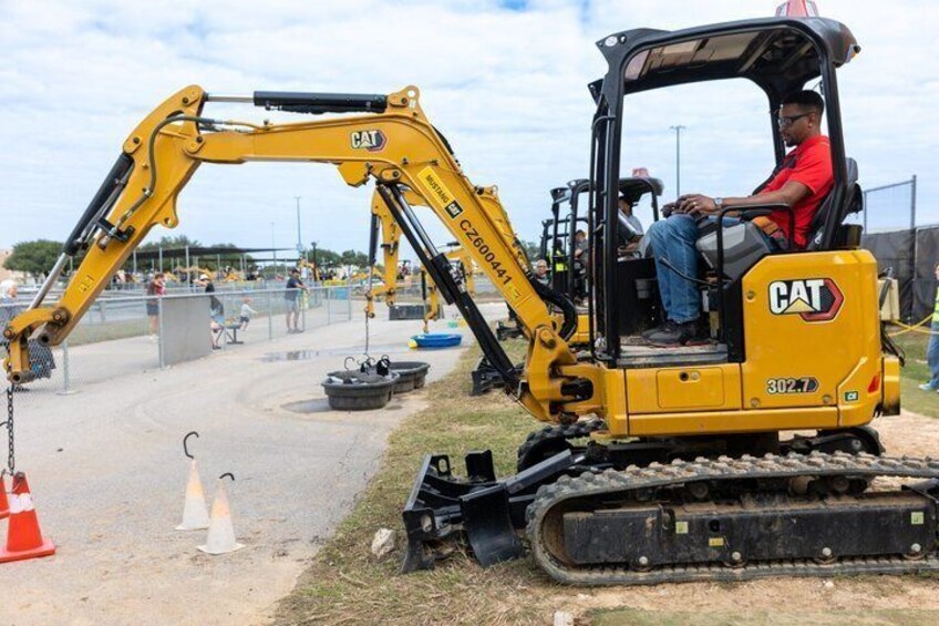 Un escavatore giallo CAT con un operatore all'interno, su un cantiere con una strada sterrata e coni stradali. 2