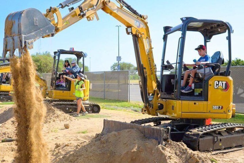 Un cantiere edile con un escavatore giallo e un veicolo giallo più piccolo, entrambi con marchio 'CAT', e una persona che opera l'escavatore indossando una camicia blu. 1