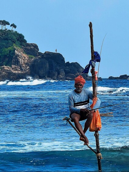 A person sitting on a wooden structure by the sea, with a rocky coastline and waves in the background. 2