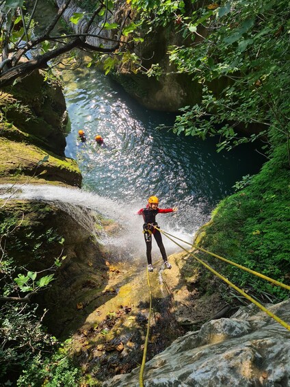 A person rappelling down a rocky cliff with a rope, surrounded by lush greenery and a body of water below. 3