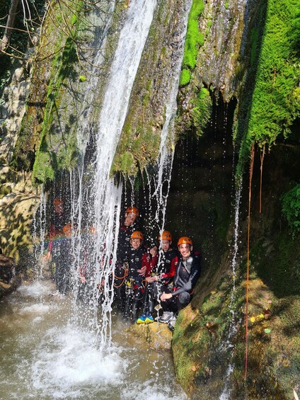 A group of people in helmets and wetsuits sitting on a moss-covered rock by a waterfall. 1