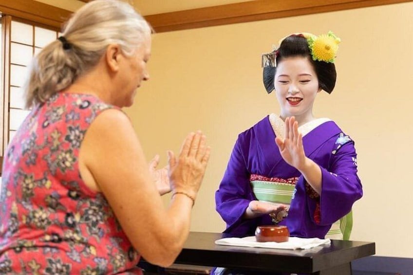A person in traditional attire with a fan and a flower accessory, seated at a table with a bowl and a small dish. 1