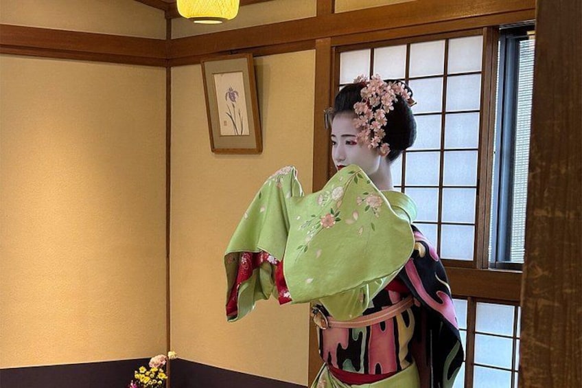 A person in traditional attire with a green kimono, holding a pink fan, standing in a room with wooden walls and a window with a grid pattern. 5
