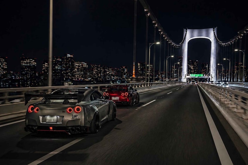 Two sports cars driving on a bridge at night with city lights in the background. 4