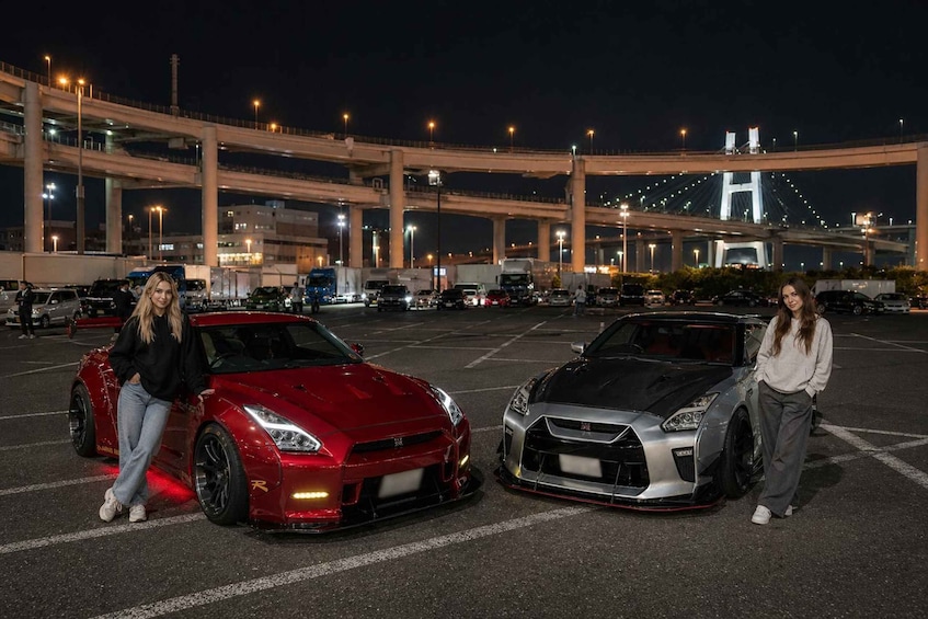 Two sports cars parked in a lot with a bridge and overpasses in the background. 5