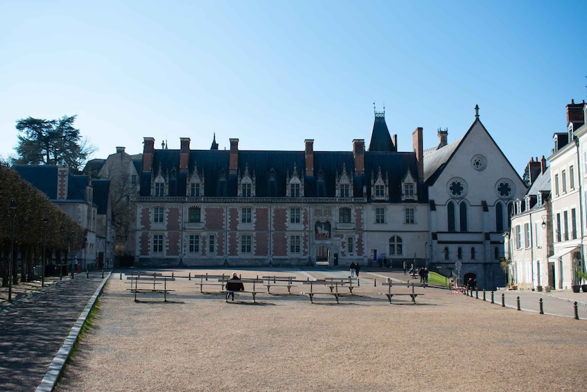 A historic building with a clock tower, a paved square with benches, and a clear sky. 1