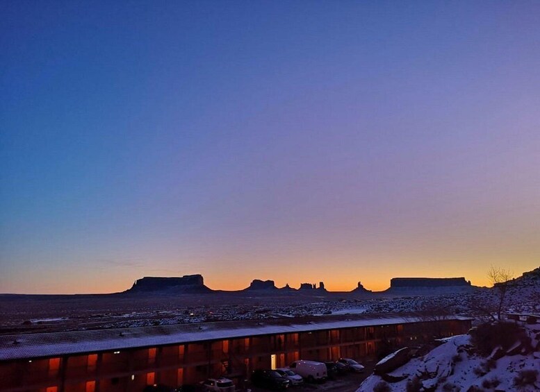 A sunset over a flat landscape with distant buttes, a parking lot with cars, and a building with red lights. 2