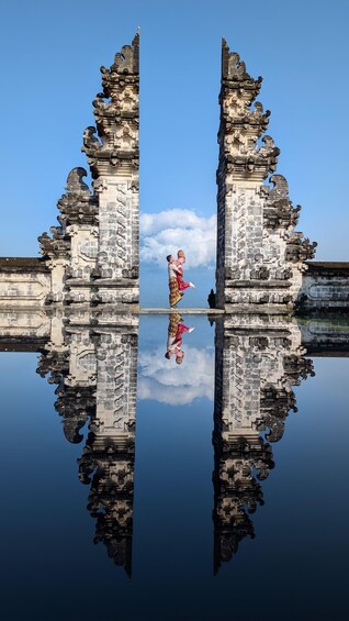 Een man en een vrouw in traditionele kleding staan op een spiegelend wateroppervlak tussen twee versierde stenen bouwwerken. 1