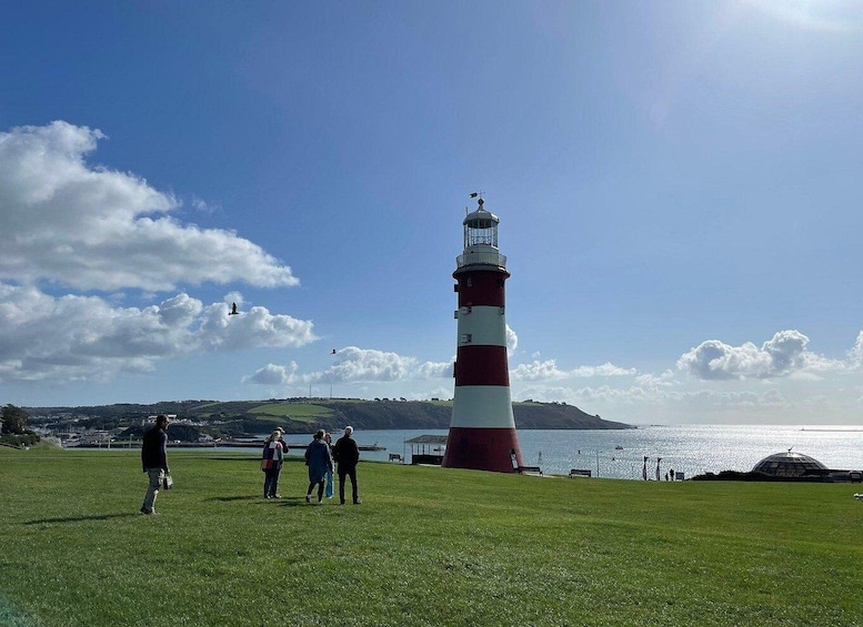 A lighthouse with people walking nearby, a grassy field, and a body of water in the background. 1