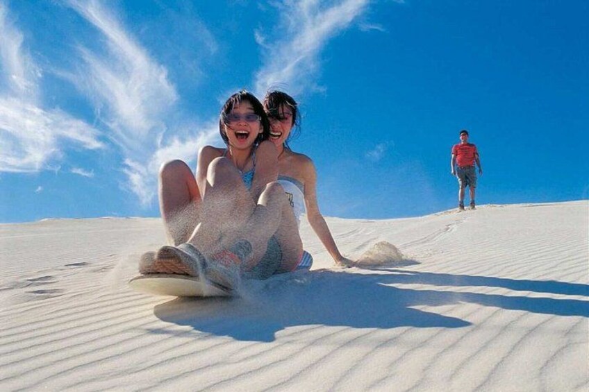 Two people sliding down a sand dune, with a third person standing in the background. 3