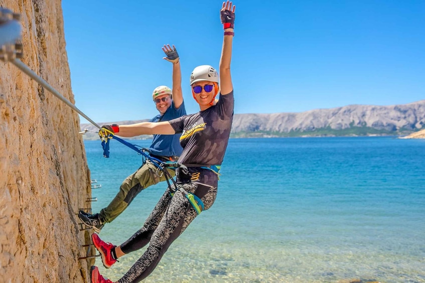 A person rappelling down a cliff with a rope, another person assisting, and a clear blue sea and hills in the background. 4